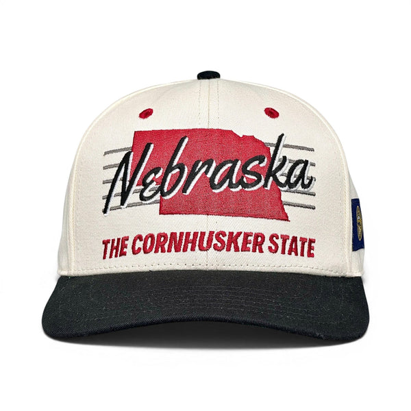 Nebraska Snapback - The Husker (Cream/Black), a retro cap featuring Nebraska's silhouette and Cornhuskers tribute, embodying vintage style and athletic heritage.
