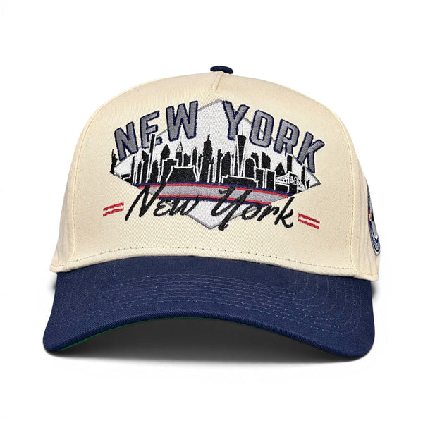 Retro New York Snapback - The Babe features a cream crown and navy brim, showcasing a vintage-style NYC skyline and New York embroidery, reminiscent of 90s Yankee Stadium nostalgia.